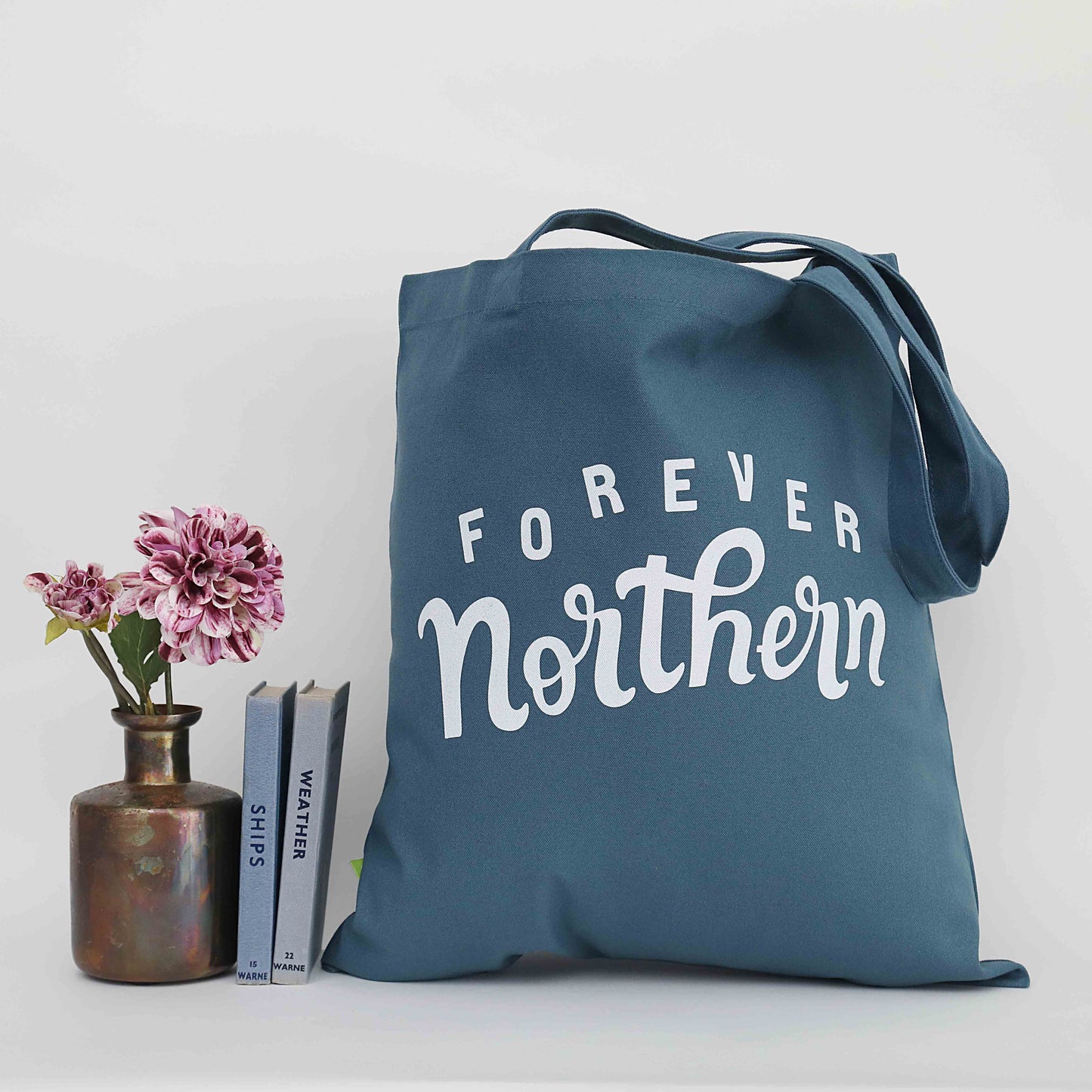 A green organic tote bag with the text 'FORE EVER NORTHERN' printed on the front, displayed alongside a vase with pink flowers and a bronze candle holder.