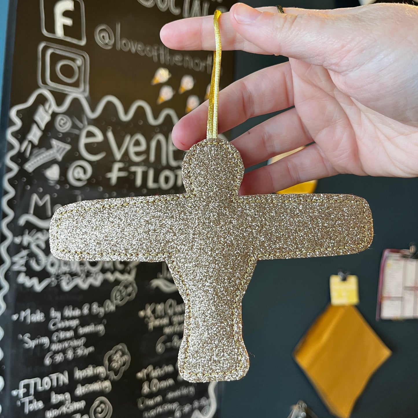 Hand holding a glittery gold angel shaped decoration on a black surface with text and logos.
