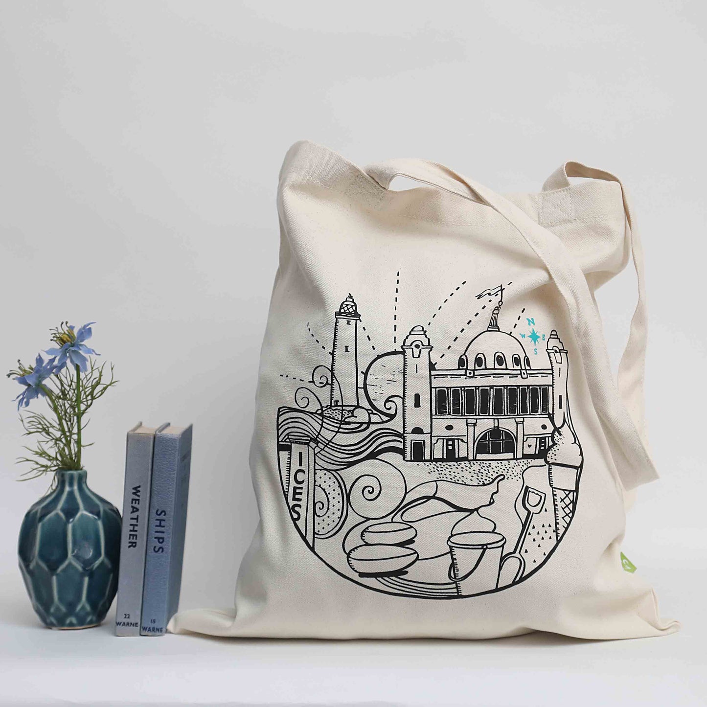 An organic cotton tote bag with a printed design featuring iconic images of places in Whitley Bay, displayed next to a vase with flowers and a book.