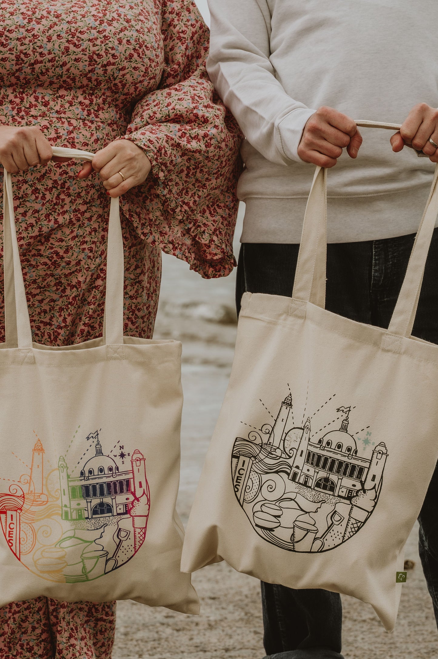 Two people holding tote bags with architectural designs on a beach.