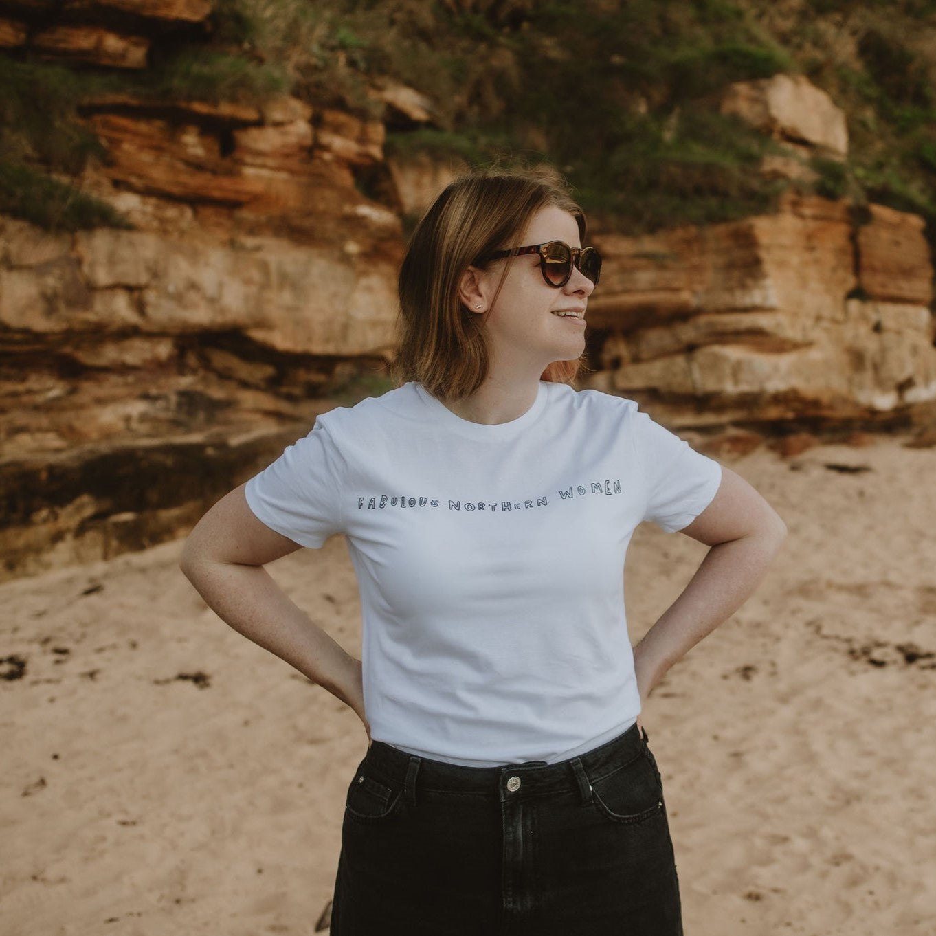 A person standing on a beach wearing a white crew neck t-shirt with text on it, and black shorts.