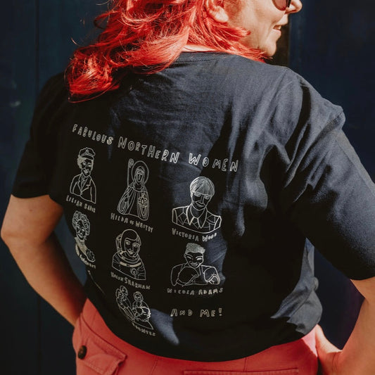 A person standing next to a dark door wearing a navy crew neck t-shirt with text and pictures of women on it, and pink trousers.
