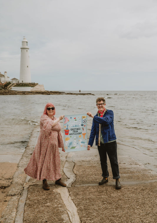 Two people holding a tea towel on a causeway with a lighthouse in the background