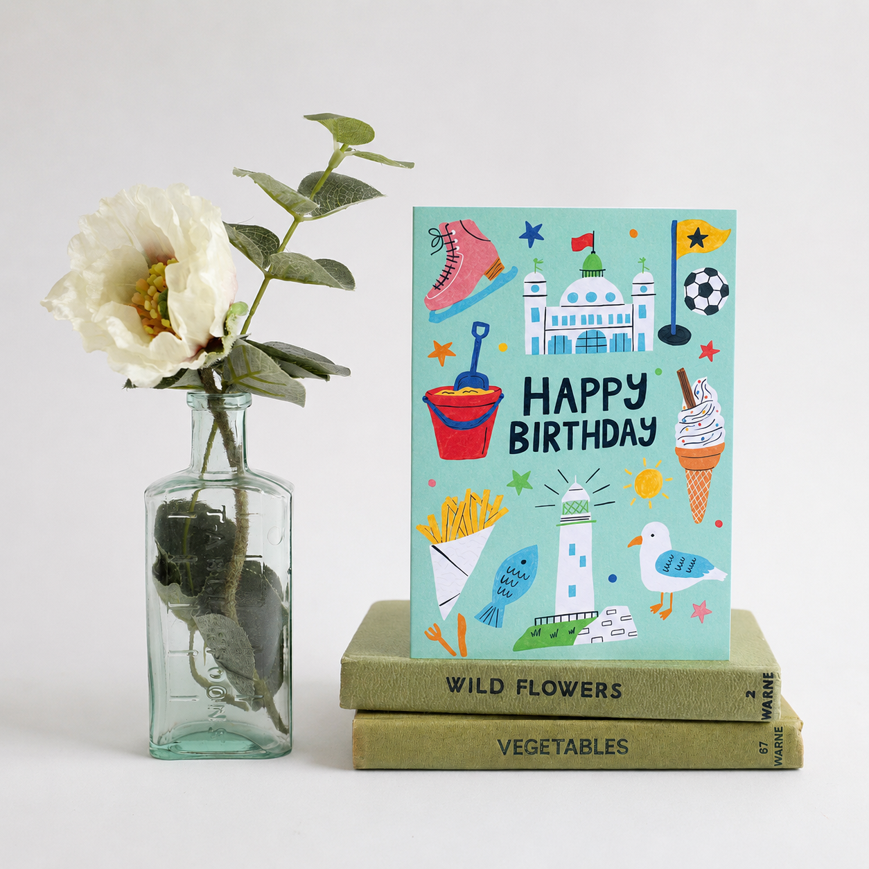 Colourful birthday card with illustrations on a stack of books next to a vase with a white flower.