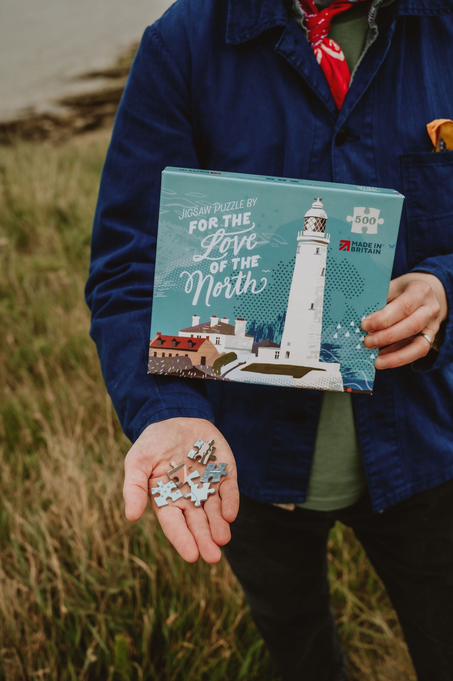Person holding a puzzle box with a lighthouse design and scattered puzzle pieces outdoors.