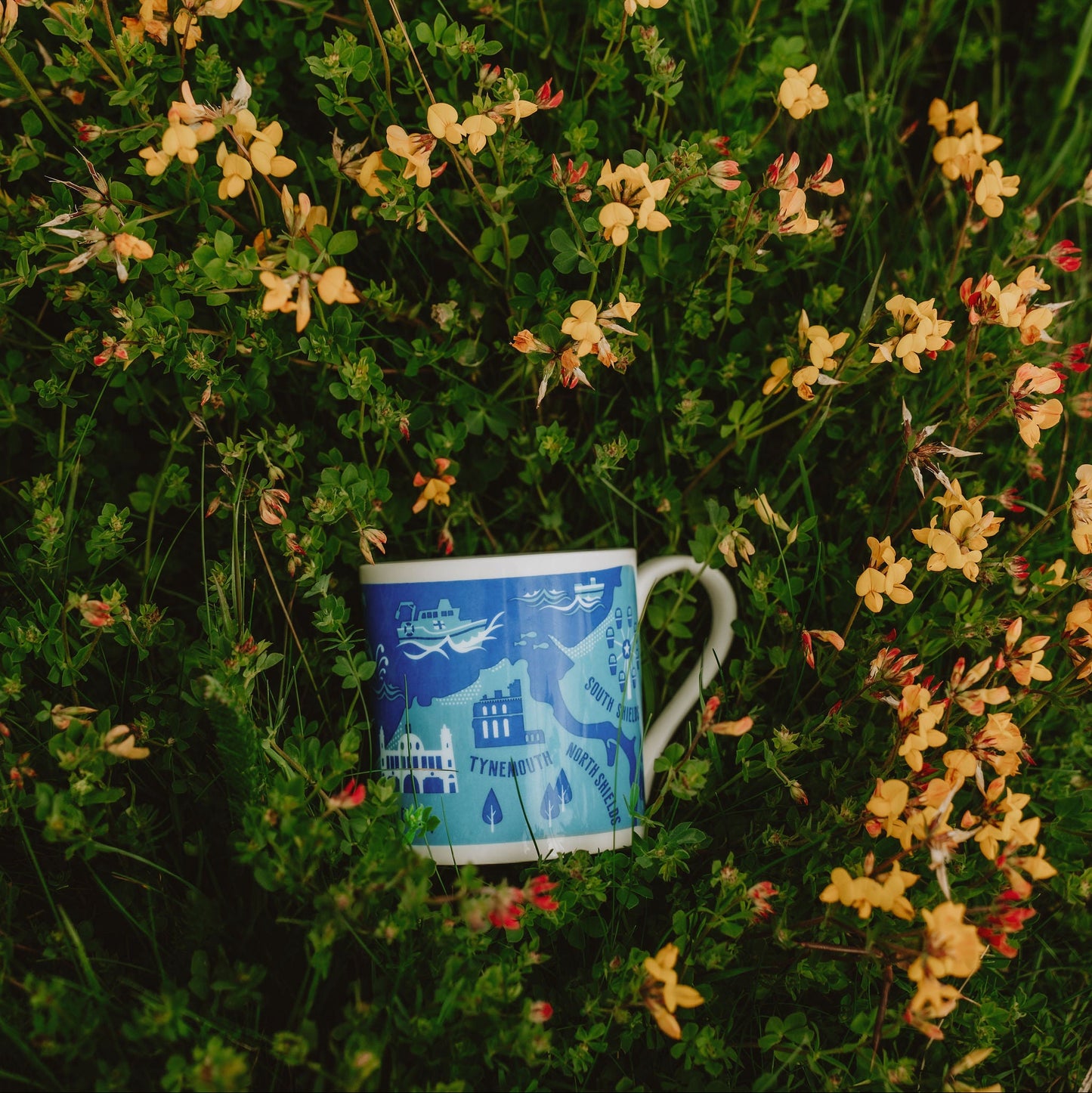Blue mug with a scenic design placed among green foliage and small yellow flowers