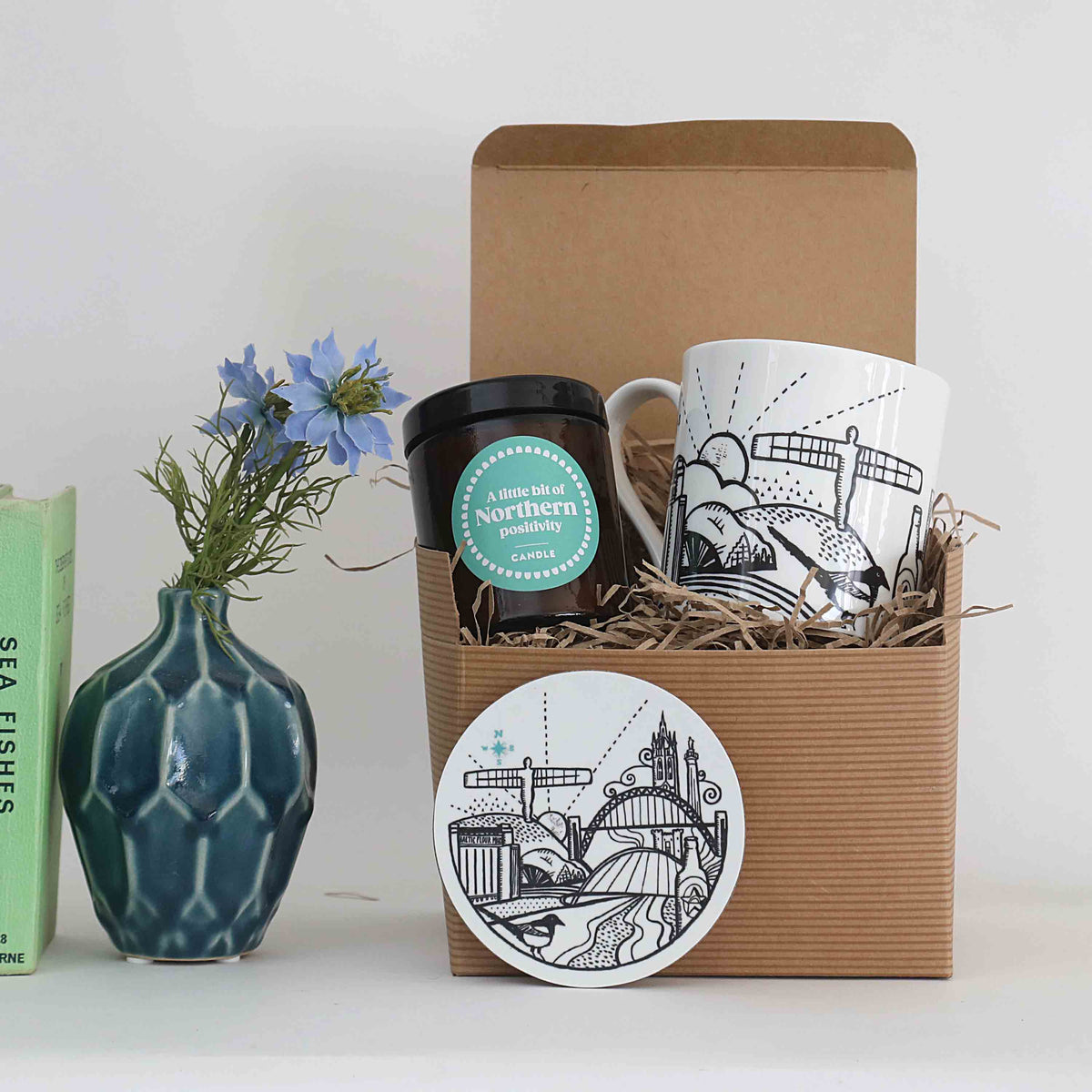 A gift set in a brown corrugated box filled with straw, containing a white ceramic mug with black illustrated artwork and a matching round coaster. A blue-green textured vase with blue flowers and a green book stand beside the box on a white surface against a light grey background.