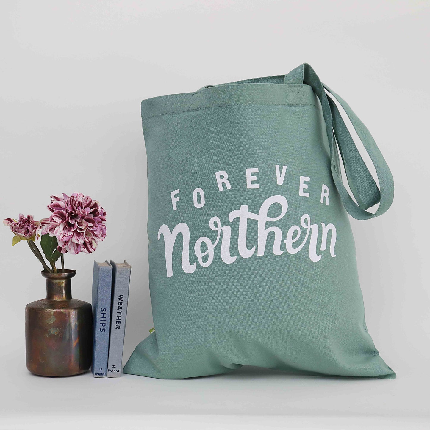 A green cotton tote bag with 'FOREVER Northern' printed in white text, displayed next to a vase with flowers and a stack of books.