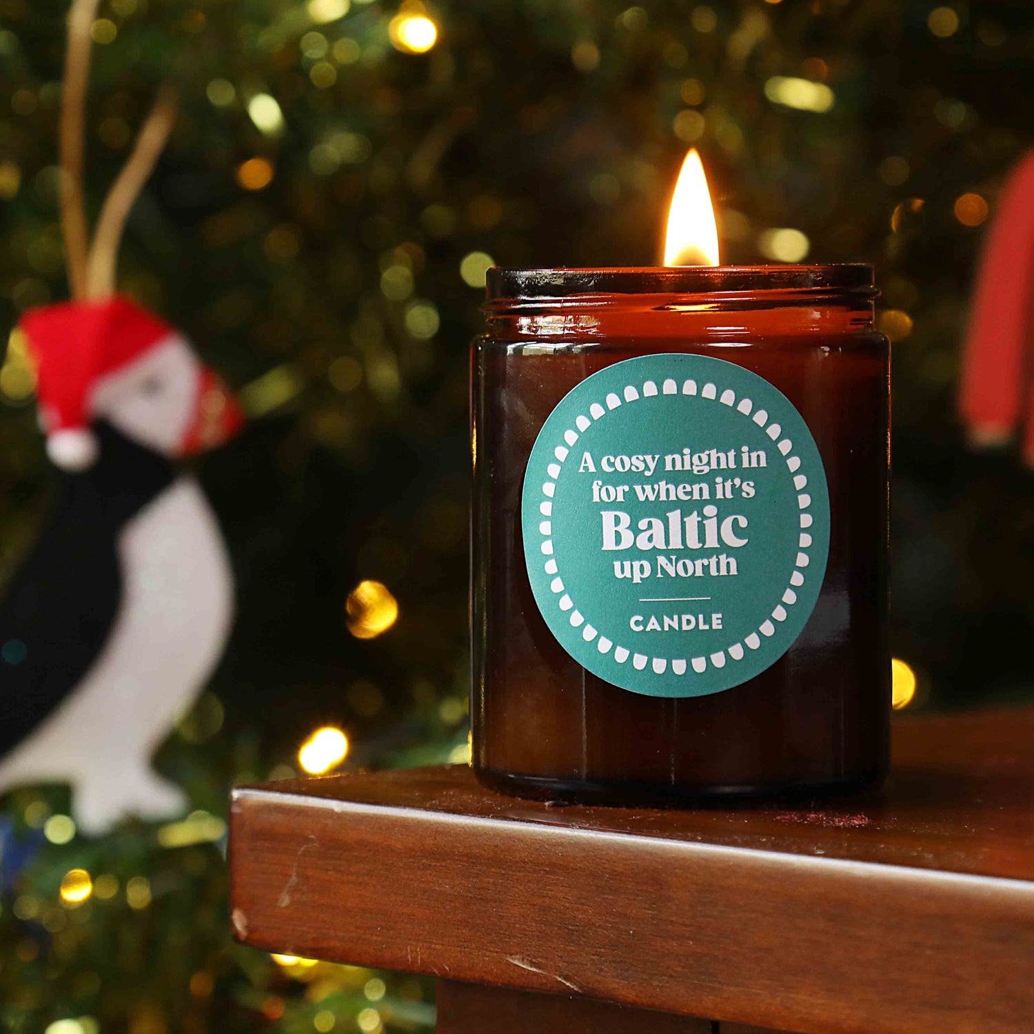A candle with a label that reads 'A Cosy Night in for when it's Baltic up North Candle', lit and placed on a wooden surface with a Christmas tree and decorations in the background.