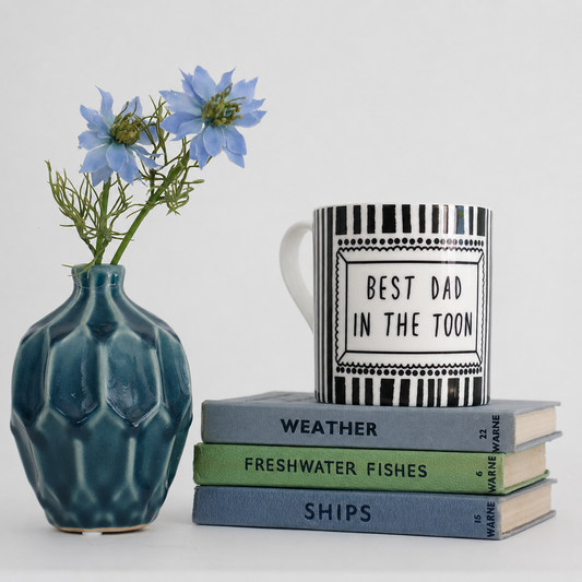 Mug with 'Best Dad in the Toon' text, stack of books, and blue vase with flowers on a light background.