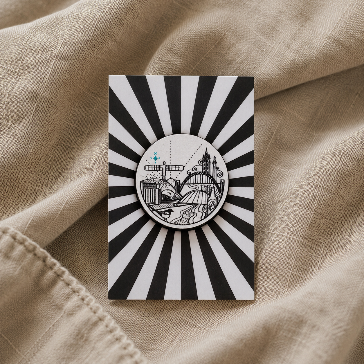 A circular black and white enamel pin featuring an illustration featuring the Angel of the North and Tyne Bridge, mounted on a matching black and white sunburst card and placed on a beige linen fabric background.
