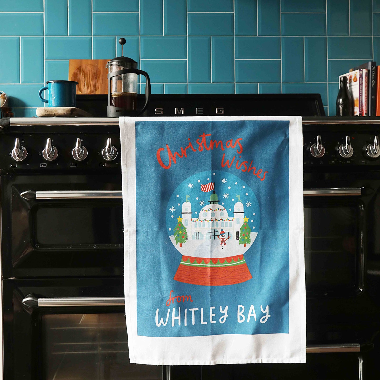 A blue organic cotton tea towel with a Christmas design, featuring the Spanish City building with festive decorations, hanging on a black range cooker.