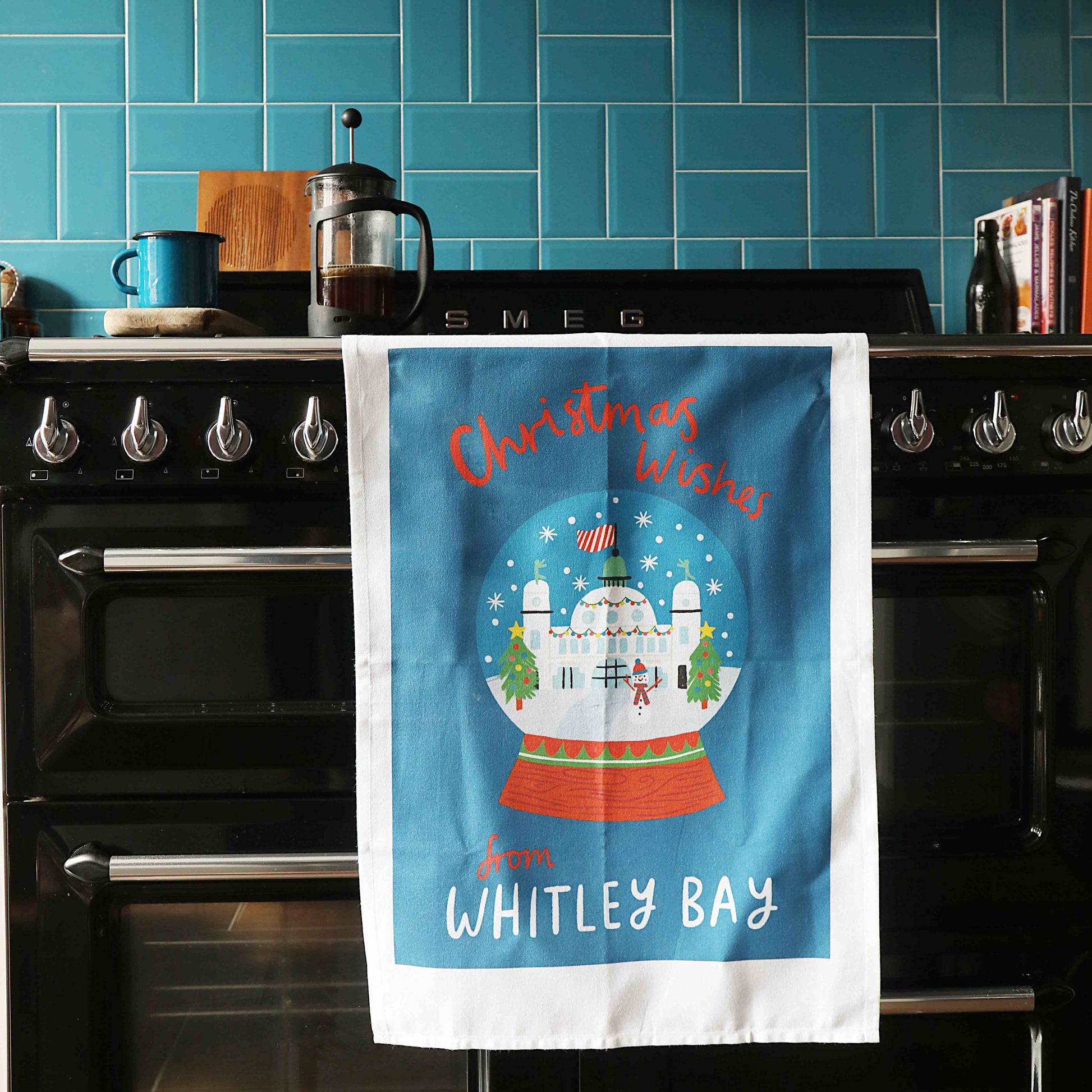 A blue organic cotton tea towel with a Christmas design, featuring the Spanish City building with festive decorations, hanging on a black range cooker.