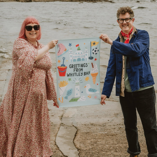 Two people holding a tea towel with 'Greetings from Whitley Bay' text and illustrations by the North Sea
