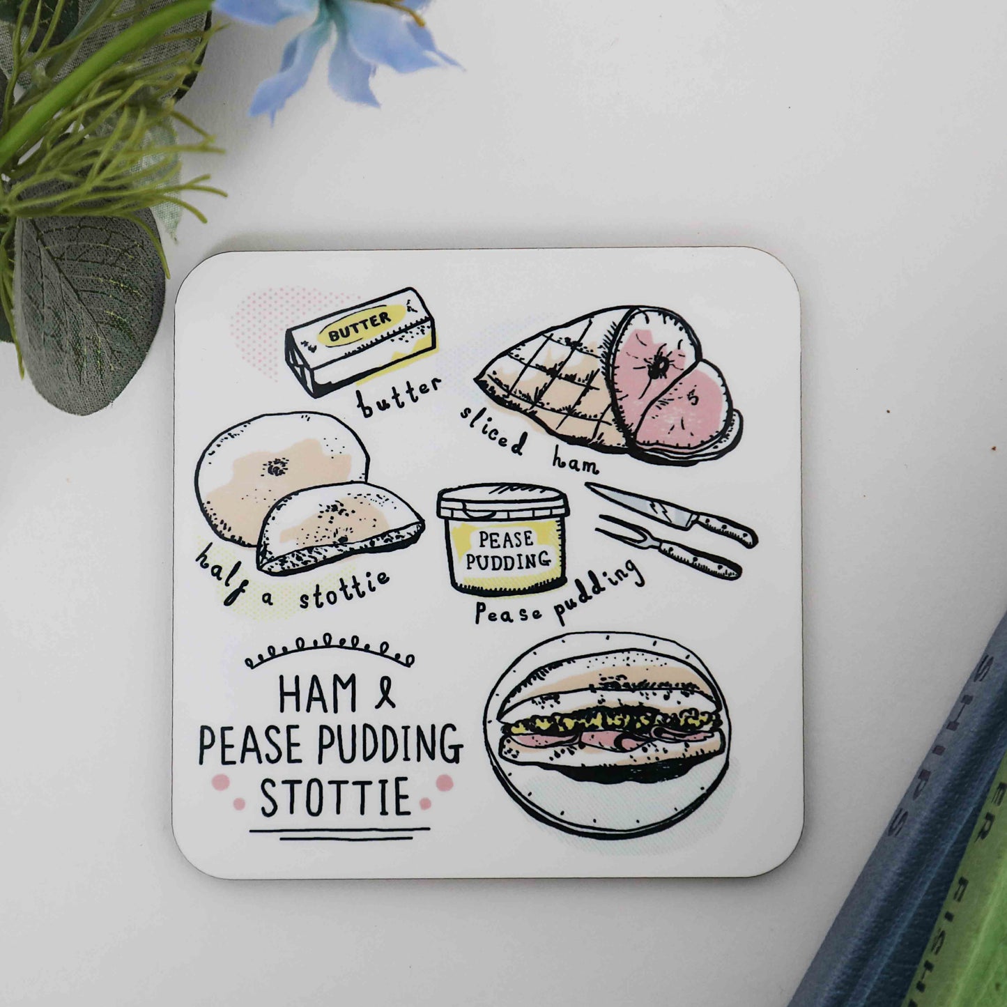 A square coaster with a cork backing, featuring an illustration of a ham and pease pudding stottie, along with other related items like a roll, butter, and a knife.