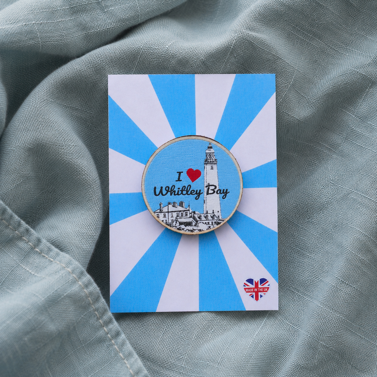 Pin with 'I ❤️ Whitley Bay' design on a blue and white striped card, placed on a textured green fabric background.