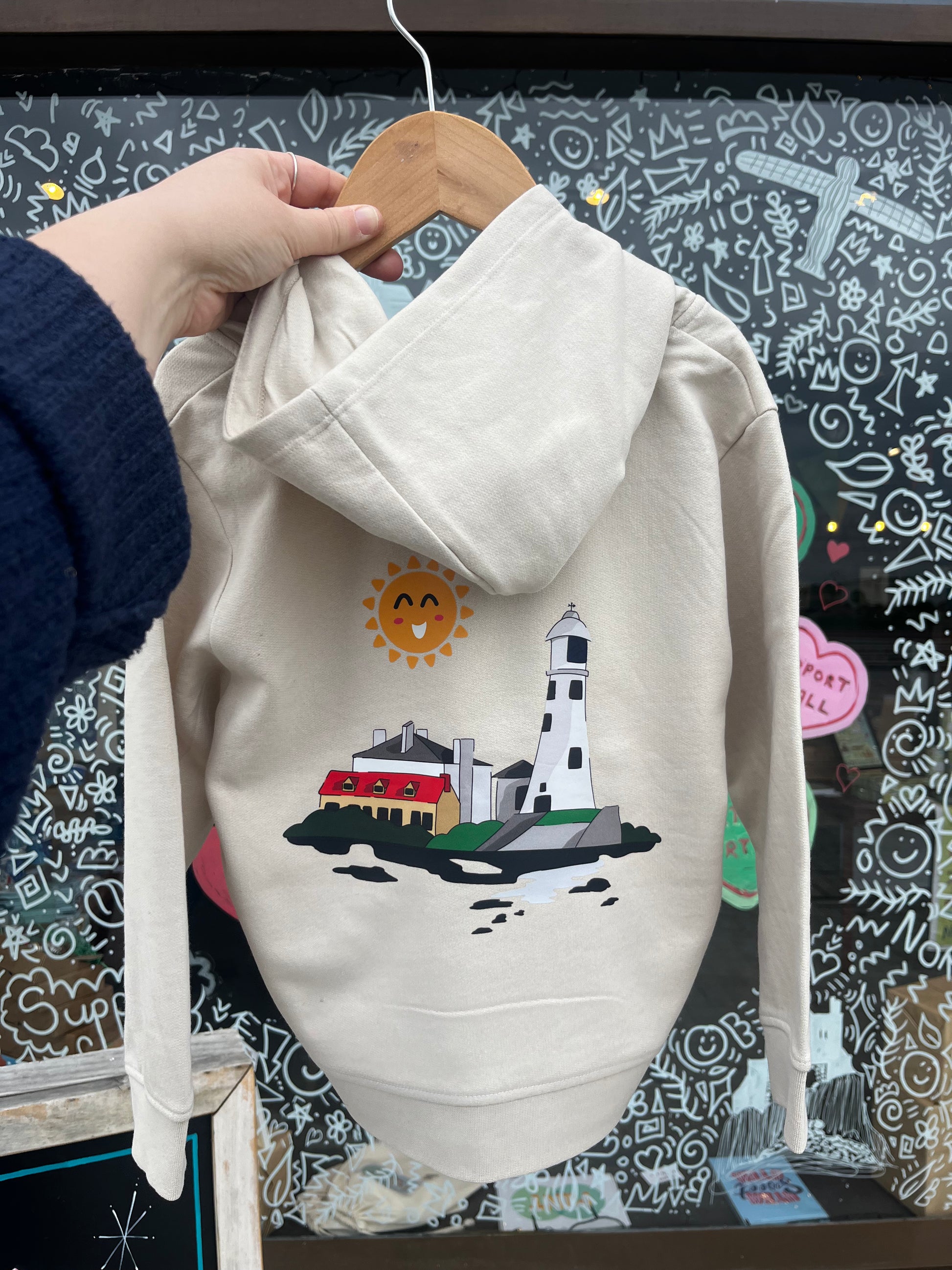 Light stone hoodie with a lighthouse design on a hanger against a chalkboard background