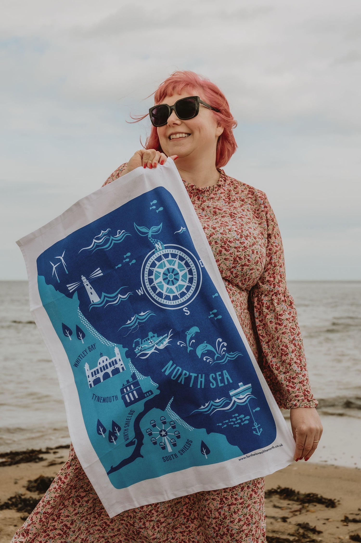 Woman holding a 'North Sea' themed tea towel on a beach.
