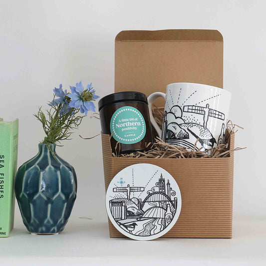 A gift set in a brown corrugated box filled with straw, containing a white ceramic mug with black illustrated artwork and a matching round coaster. A blue-green textured vase with blue flowers and a green book stand beside the box on a white surface against a light grey background.