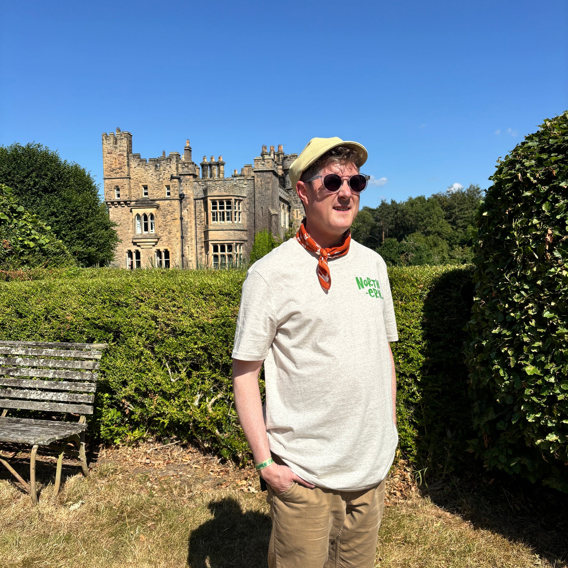 A man wearing a beige cap, sunglasses, an orange neck scarf, and a light beige t-shirt with green 'North-ern' branding stands in a garden in front of a large stone castle under a clear blue sky.