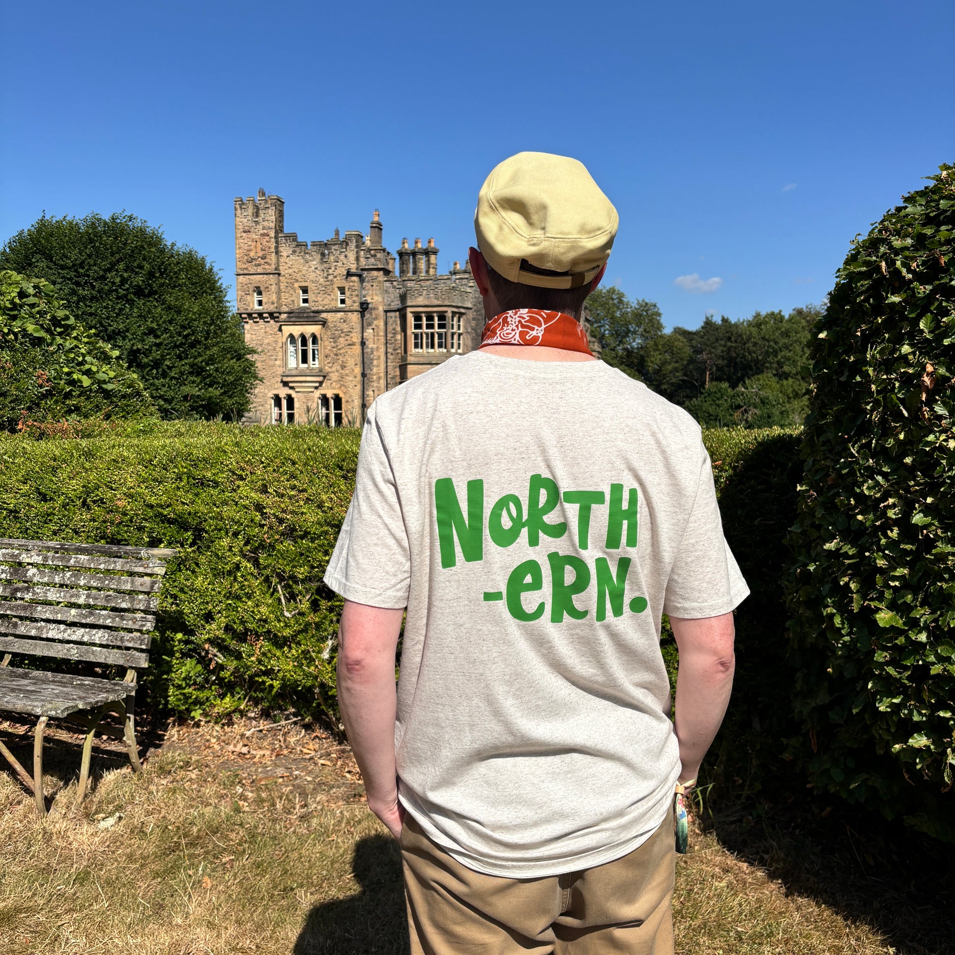 A man viewed from behind wearing a light beige t-shirt with 'NORTH -ERN.' printed in green across the back, standing in a garden with hedges and a stone castle in the background under a blue sky.