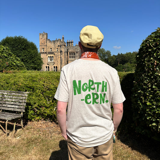 A man viewed from behind wearing a light beige t-shirt with 'NORTH -ERN.' printed in green across the back, standing in a garden with hedges and a stone castle in the background under a blue sky.