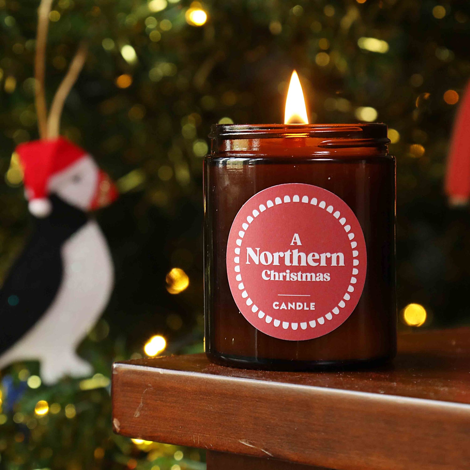 A lit candle with a red label that reads 'A Northern Christmas CANDLE', placed on a wooden surface with a blurred Christmas tree and decorations in the background.