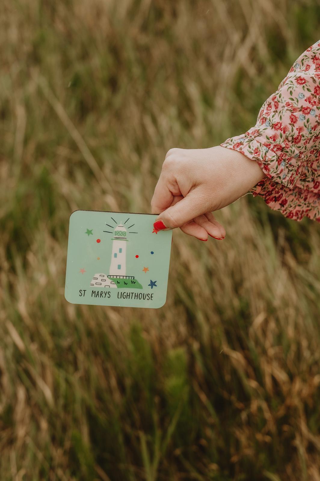 Hand holding a card with a lighthouse coaster in a grassy field.