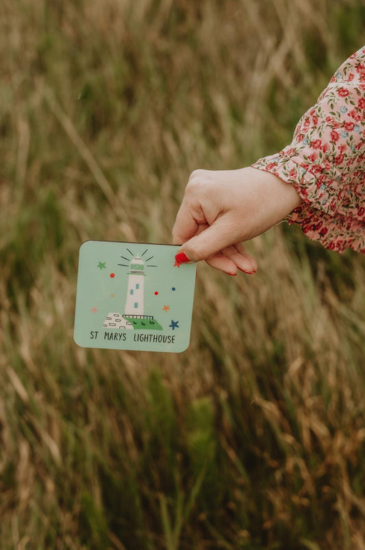 Hand holding a card with a lighthouse coaster in a grassy field.