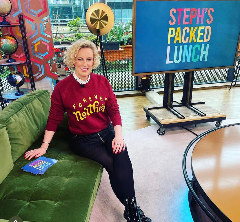 A person sitting on a green sofa wearing a red sweatshirt with text, in front of a sign that reads 'STEPH'S PACKED LUNCH'.
