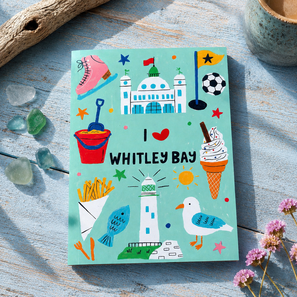 Notebook with colourful illustrations of Whitley Bay on a wooden surface with flowers and driftwood.