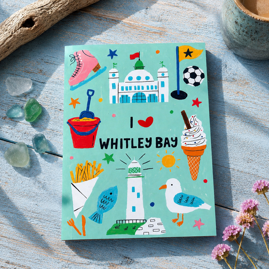Notebook with colorful illustrations of Whitley Bay on a wooden surface with flowers and driftwood.