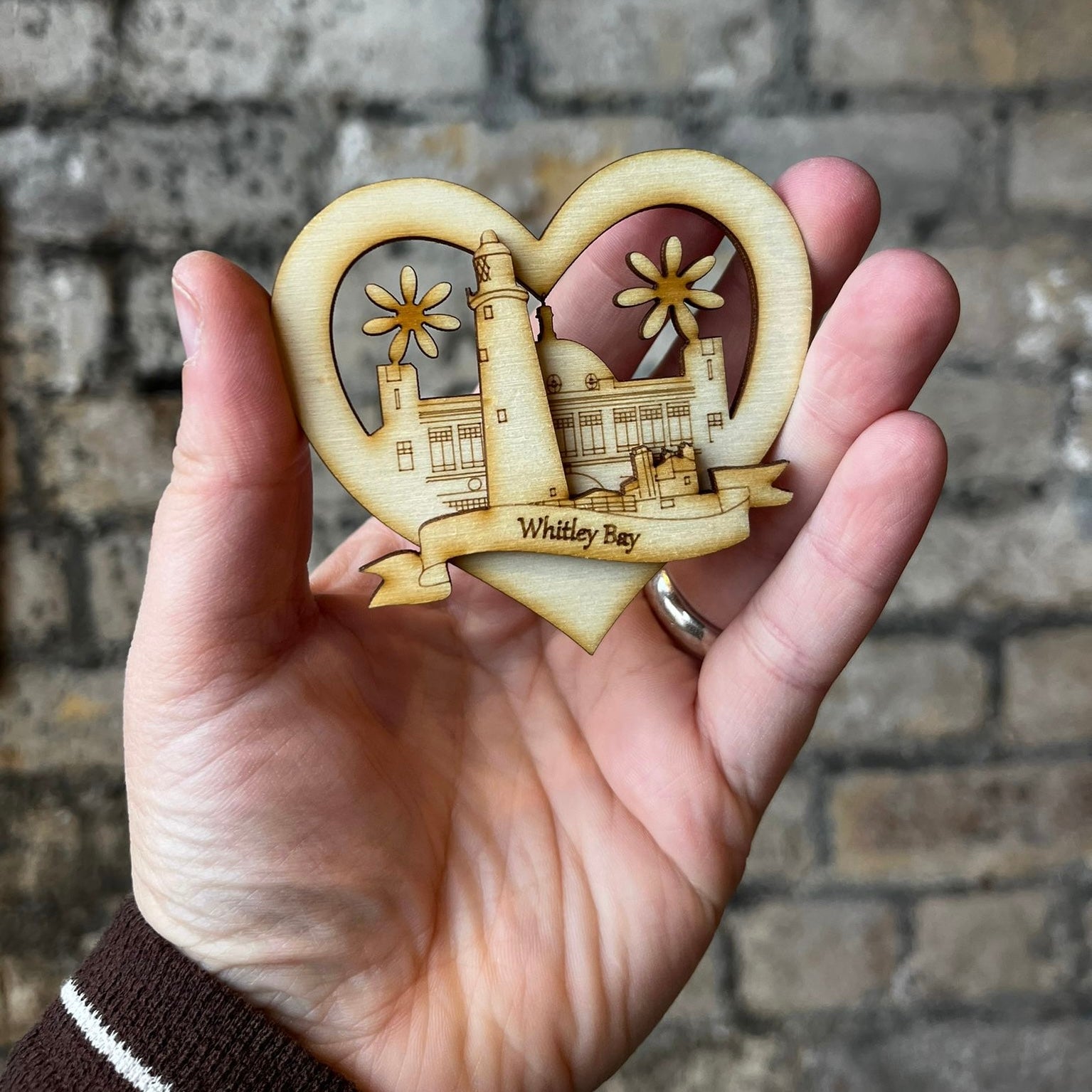 Heart-shaped ornament with a lighthouse design held in a hand against a brick wall.