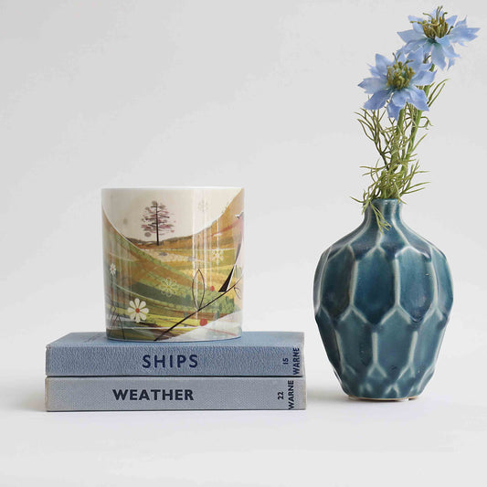 A mug with a landscape depiction, placed on top of two books titled 'SHIPS' and 'WEATHER', next to a blue patterned vase with flowers.