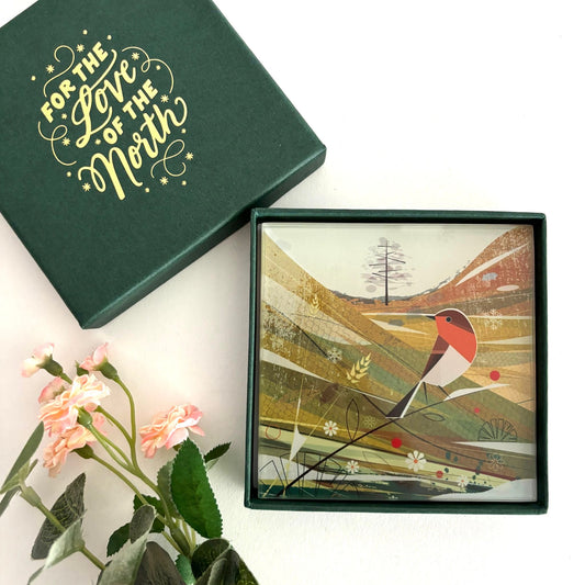 A glass coaster with a winter scene featuring a bird and sycamore trees, presented in a green gift box with the text 'For the love of the North'.