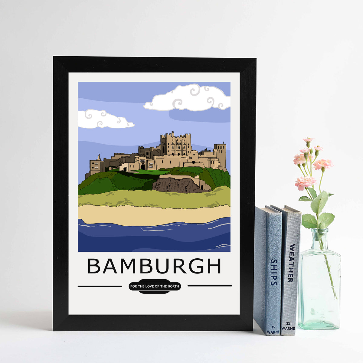 A framed print of Bamburgh featuring a vintage style illustration of the castle and beach, placed on a shelf with a vase of flowers and books.