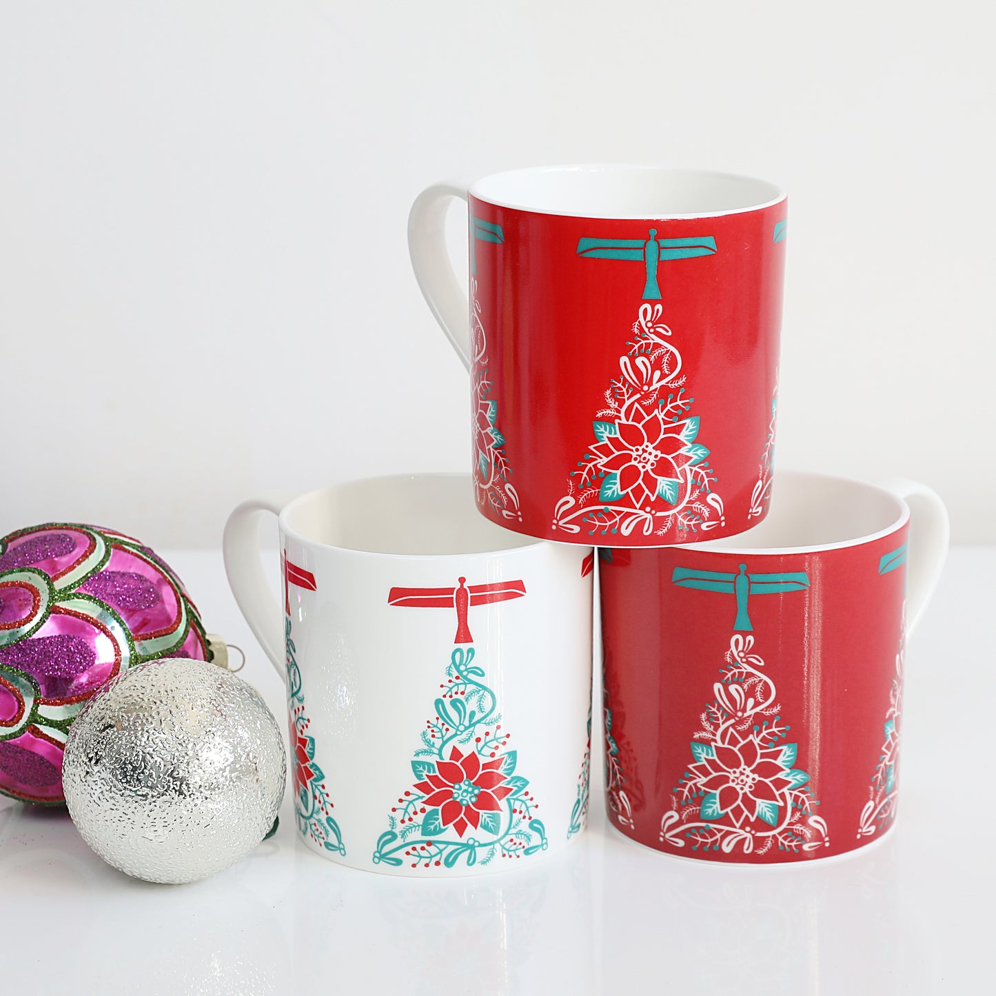 A set of three Christmas-themed china mugs with a red and white design featuring an angel and a Christmas tree, displayed stacked with one mug in front of the other on a white surface, with a decorative ball to the side.