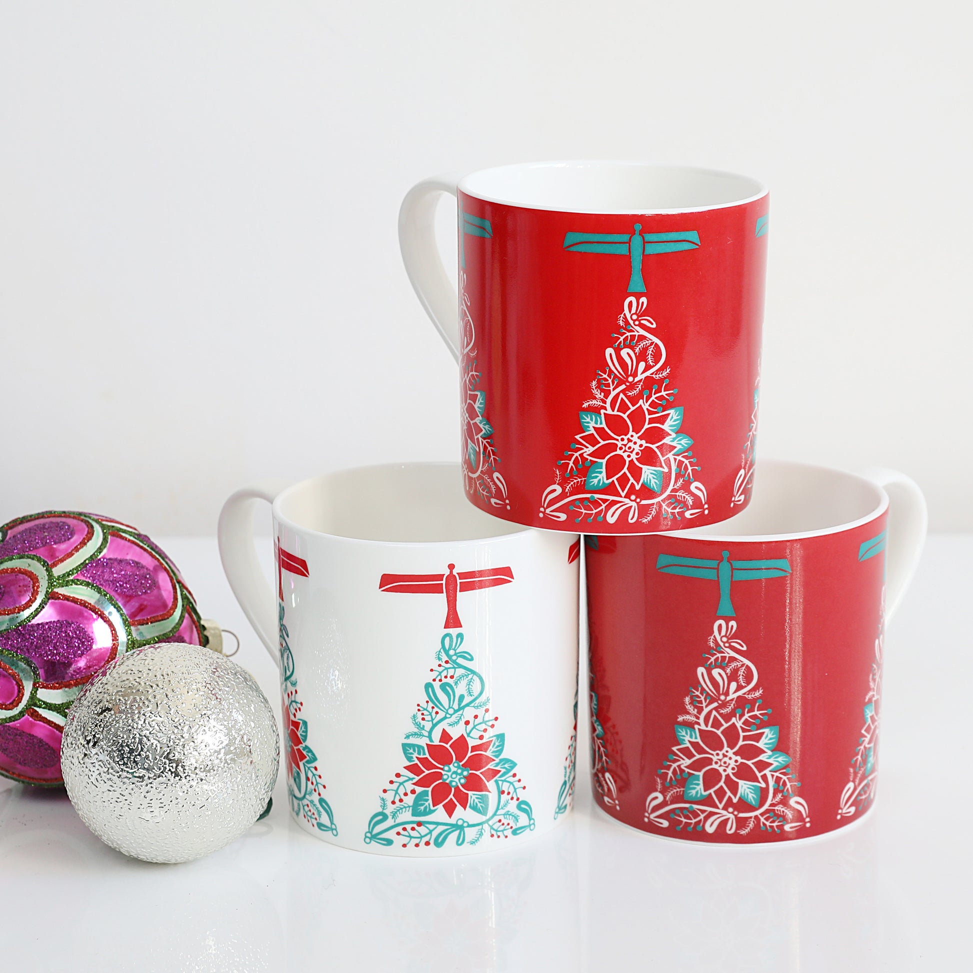 A set of three Christmas-themed china mugs with a red and white design featuring an angel and a Christmas tree, displayed stacked with one mug in front of the other on a white surface, with a decorative ball to the side.