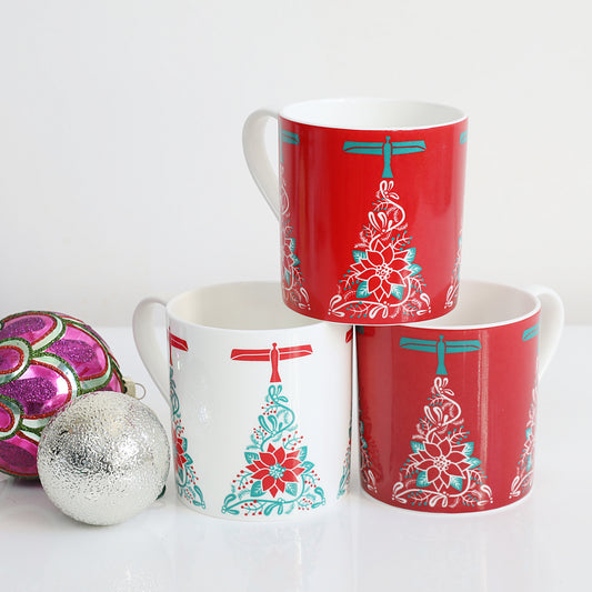 A set of three Christmas-themed china mugs with a red and white design featuring an angel and a Christmas tree, displayed stacked with one mug in front of the other on a white surface, with a decorative ball to the side.