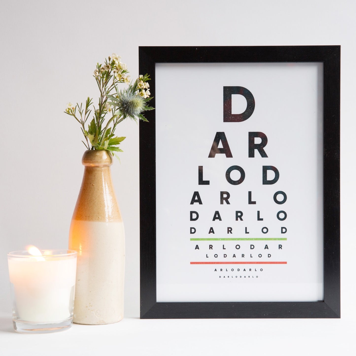 A framed print with the text 'DARLO DARLO DARLO DARLO' in bold, graphic lettering, displayed on a white surface with a vase of flowers and a candle to the side.
