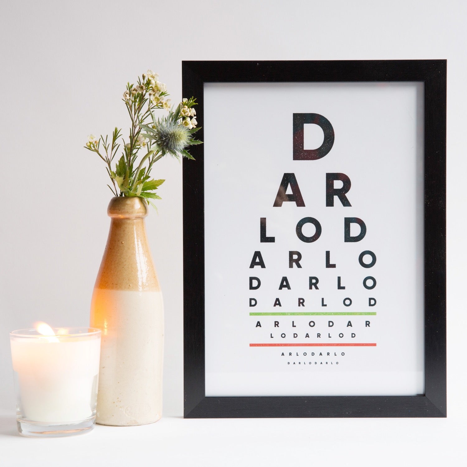 A framed print with the text 'DARLO DARLO DARLO DARLO' in bold, graphic lettering, displayed on a white surface with a vase of flowers and a candle to the side.