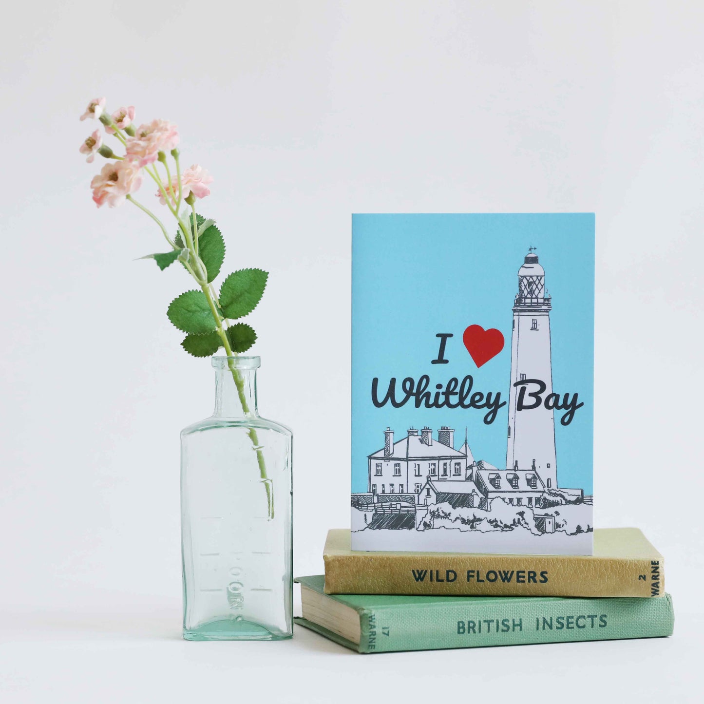 A greeting card with a blue background featuring an illustration of St Mary's Lighthouse in Whitley Bay, accompanied by a small bouquet of pink flowers and placed on a surface with several books and a glass vase with flowers.