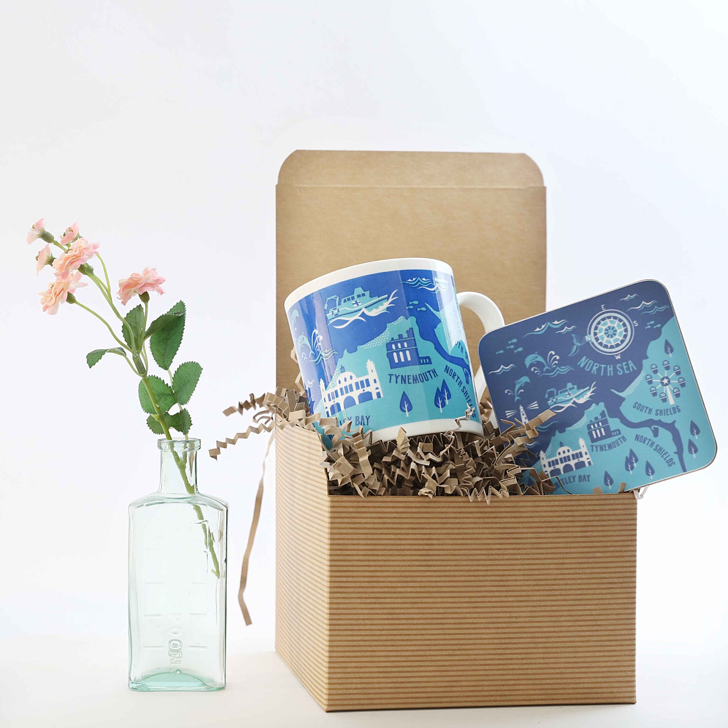 A gift box open to reveal a blue and white mug with a cityscape design, a matching coaster, and crumpled paper filling. To the left, there is a clear glass vase with flowers inside it.
