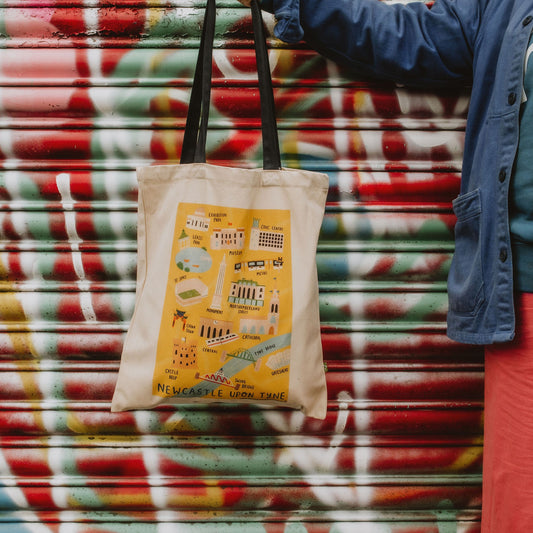 A tote bag with a colorful pattern featuring iconic Newcastle locations, displayed against a patterned background.