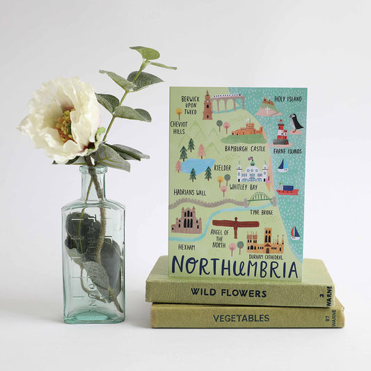 A greeting card depicting an illustrated map of Northumbria, with iconic landscapes and landmarks, placed on top of two books with a vase of flowers to the left.